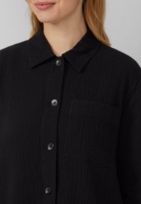 Schwarze Button-Up-Bluse aus strukturiertem Stoff, mit klassischem Kragen, Brusttasche und fünf passenden Knöpfen an der Knopfleiste.