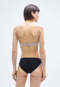 Ανοιχτό γκρι σουτι έν με λεπτές ρυθμιζόμενες τιράντες και κούμπωμα, σε συνδυασμό με μαύρο σλιπ χαμηλού καβάλου, εμφανιζόμενα από πίσω σε ουδέτερο φόντο.