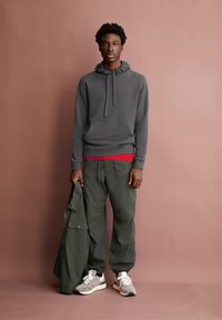 Felpa grigia con cappuccio e scollo arricciato, maglietta rossa, pantaloni cargo verdi oliva e sneakers grigie, mentre tiene in mano una giacca verde.