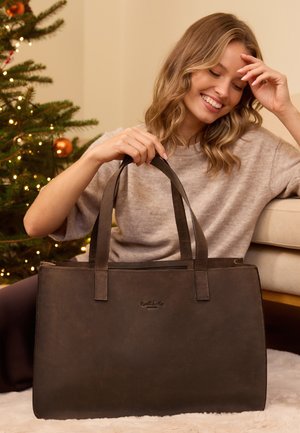 Donna sorridente seduta sul pavimento vicino all'albero di Natale decorato che tiene una grande borsa a tracolla marrone in pelle con doppi manici.