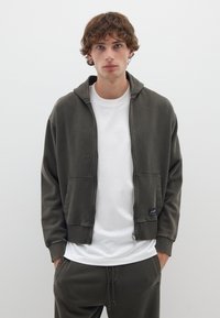 Felpa con zip verde oliva, dotata di tasche frontali, polsini a coste e un logo. Indossata sopra una maglietta bianca e pantaloni della tuta coordinati in verde oliva.