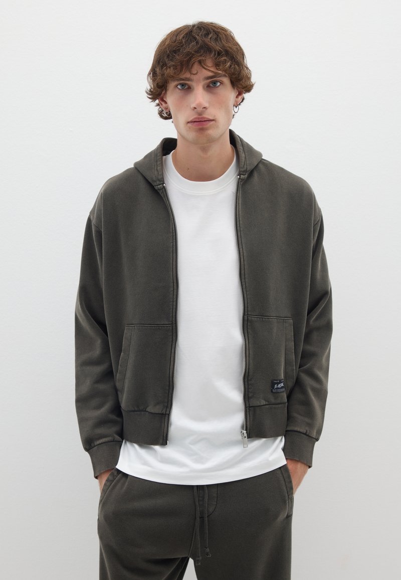 Felpa con zip verde oliva, dotata di tasche frontali, polsini a coste e un logo. Indossata sopra una maglietta bianca e pantaloni della tuta coordinati in verde oliva.