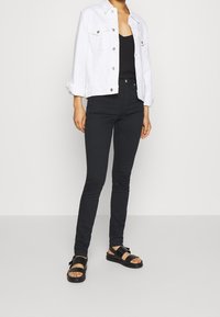 Valkoinen farkkutakki, jossa on nepparit, musta toppi, mustat kiiltävät farkut ja mustat sandaalit paksuilla remmeillä ja tasaisella pohjalla.