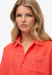 Koralowa, teksturowana koszula z tłoczonym wzorem sieci, zapinana na guziki z kieszenią na piersi po lewej stronie z wyhaftowanym logo „Superdry”.