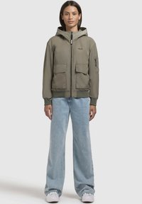 Doudoune verte olive avec capuche, fermeture éclair à l'avant et poignets élastiques. Dispose de deux poches avant et d'une poche sur le bras. Associée à un jean en denim à jambes larges.