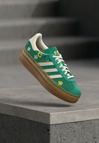 Zapatilla verde con diseños florales, tres rayas blancas y una suela gruesa de goma. Cuenta con una parte superior texturizada y cordones blancos. Etiqueta de la marca en la lengüeta.