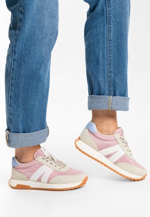 Pieds chaussés de baskets roses, beiges et blanches avec des semelles orange, associés à un jean bleu retroussé sur fond blanc.