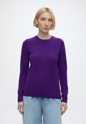 Woman with short blonde hair wearing a purple long-sleeve sweater and light blue jeans, standing against a plain white background.
