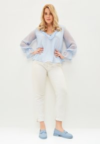 Lichtblauwe blouse met ruches rond de halslijn en transparante mouwen, gecombineerd met witte opgerolde jeans en lichtblauwe instappers.