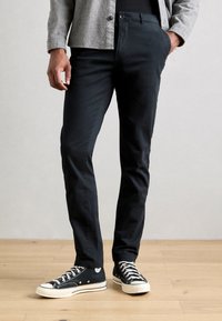 Un pantalon noir en coton présente une coupe slim avec une texture lisse. Associé à des baskets noires montantes et un cardigan gris pour le contraste.