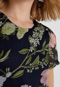 Námořnická květinová blůza s krátkým rukávem, s zelenými a růžovými květy na texturované tkanině. Kulatý výstřih a jemný lesk.