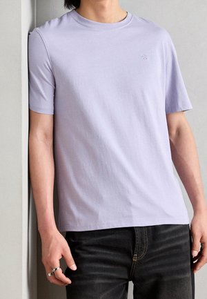 T-shirt à manches courtes couleur lavande claire en tissu doux, avec un col rond et un petit logo brodé sur la poitrine.