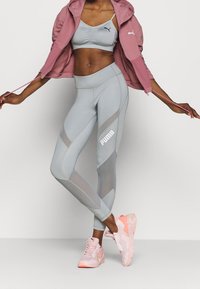 Tenue de sport gris clair comprenant un brassière et un legging taille haute, avec des accents en mailles et un logo Puma bien visible. Associée avec des baskets rose pâle.