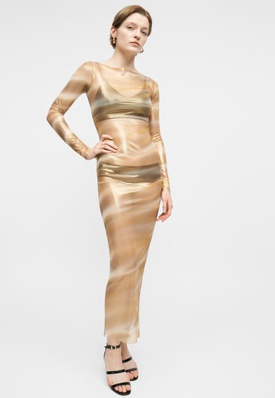 Γυναίκα που φοράει λαμπερό, εφαρμοστό, μακρυμάνικο, μακρύ έως το πάτωμα φόρεμα σε χρυσό και μπεζ χρώμα με μαύρα πέδιλα με λουριά, ποζάροντας με το ένα χέρι στη μέση.