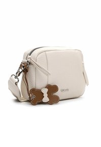 Kremowa skórzana torba na ramię z zamkiem błyskawicznym, o zaokrąglonym kształcie i brązowym zawieszce w kształcie kości. Teksturowana powierzchnia z subtelnym połyskiem.