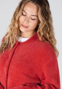 Rode, zacht aanvoelende jacket met een ronde halslijn en knoopsluiting. De stof lijkt pluizig, wat warmte en comfort biedt.