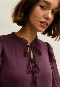 Vrouw met kort bruin haar, die een pruimkleurige blouse draagt met een gerimpelde kraag en een striksluiting aan de voorkant, tegen een neutrale achtergrond.