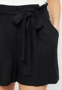 Μαύρο σορτς με ψηλή μέση και υφασμάτινη ζώνη δεμένη σε φιόγκο, πλαϊνές τσέπες και κοντινό πλάνο ενός χεριού που ακουμπά σε μία από τις τσέπες.