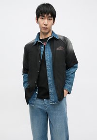 Farmere és fekete szövetből készült ing, gombos, kontrasztos rétegekkel. Az egyik oldalon fekete panel, a másikon farmer található, finom logó részlettel.