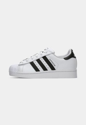 Witte Adidas Superstar sneaker met zwarte strepen, schelpneus en de tekst "Superstar" op de zijkant, weergegeven in linkse profiel op een witte achtergrond.