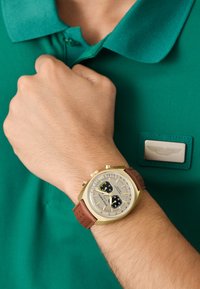 Chronografické hodinky s puzdrom v zlatej farbe, béžovým ciferníkom, dvoma čiernymi subciferníkmi a hnedým koženým remienkom, nosené s zeleným polo tričkom.