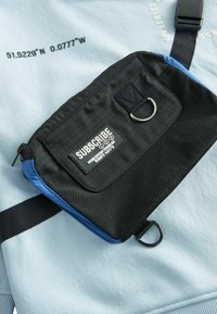 Gaiši zils audums ar melnu un zilu sietiņu, kas satur zīmola nosaukumu, rāvējslēdzēju, cilpu un melnā krāsā drukātas koordinātes.