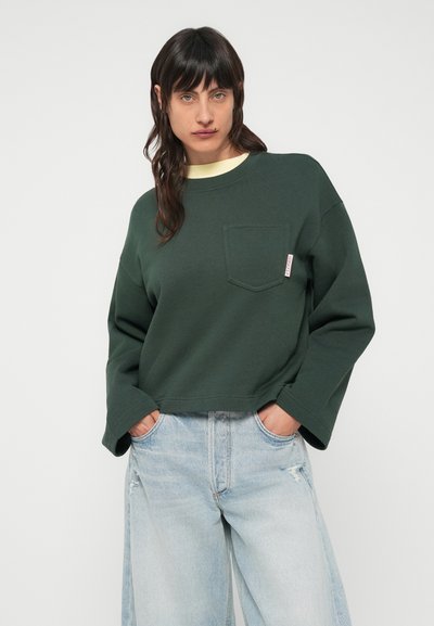 Πράσινο κοντό φούτερ με τσέπη, φαρδιά μανίκια και αντίθετο κίτρινο εσωτερικό γιακά, συνδυασμένο με ανοιχτό μπλε τζιν με φθορές.