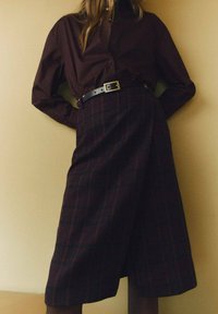 Camisa de botones marrón oscuro combinada con una falda midi de cuadros en tonos oscuros, ceñida a la cintura con un cinturón de cuero negro.