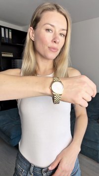 Kuldkell, millel on ümmargune nägu ja tekstuuriga metallist rihm, kantud heledanahaliselt randmelt. Taustal on tume riiul ja sinine diivan.