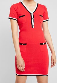 Robe en maille rouge à manches courtes, col en V, finitions blanches et noires, deux poches avant, et détails de boutons sur la patte de boutonnage. Texture lisse.