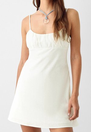 Kvinde iført hvid, ærmeløs minikjole med rynket brystparti og en blå halskæde med et rundt gennemsigtigt vedhæng.