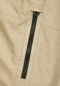 Beige tyg med en vertikal svart dragkedja, med texturerade tänder och en liten rund dragflik. Inkluderar en sidoficka.