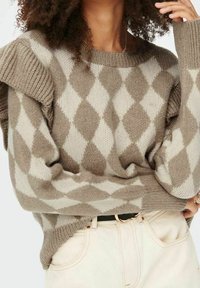 Pull à motif losanges marron et beige avec texture côtelée, doté de détails à volants aux épaules et de manches longues, porté sur un pantalon clair.