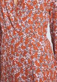 Blommigt mönstrad omslagsklänning i orangea och rosa nyanser. Har en mjuk, lätt vikt och en draperad midja samt långa ärmar.