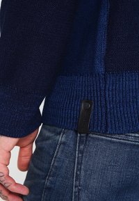 Marinblå stickad tröja med ribbstickade muddar och kant; har ett texturerat mönster, svart etikett fäst vid fållen, bärs tillsammans med denimjeans.