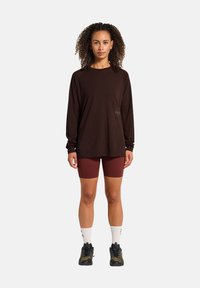 Langärmliges, dunkelbraunes Shirt mit einem entspannten Schnitt und Logodetail. Kombiniert mit burgunderfarbenen Radshorts und schwarzen Sneakers mit grünen Akzenten.