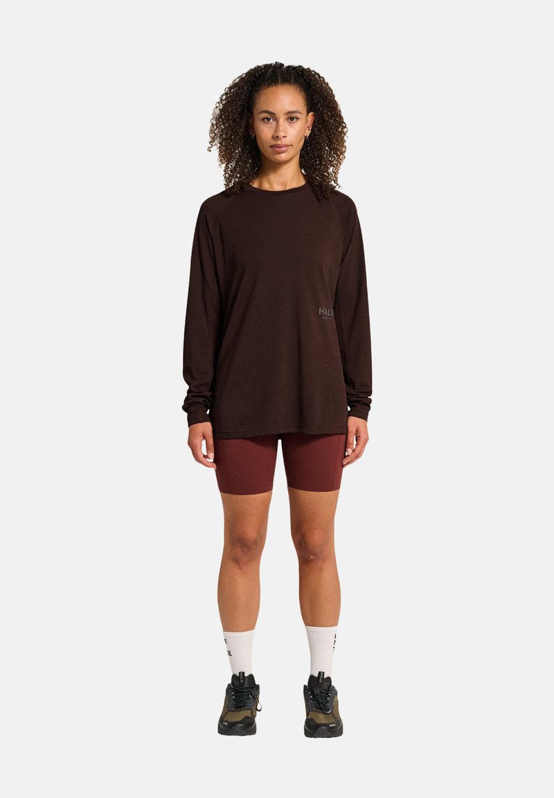 Langärmliges, dunkelbraunes Shirt mit einem entspannten Schnitt und Logodetail. Kombiniert mit burgunderfarbenen Radshorts und schwarzen Sneakers mit grünen Akzenten.