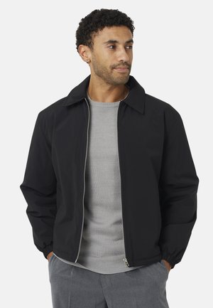 Hombre con cabello corto y rizado que lleva una chaqueta negra con cremallera encima de un suéter gris claro y pantalones grises, mirando hacia su derecha con las manos en los bolsillos.