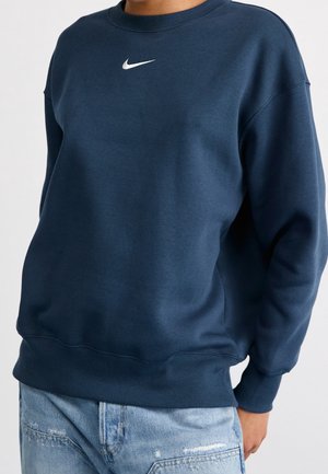 Osoba ubrana w granatową bluzę Nike z białym logo Swoosh, w połączeniu z jasnoniebieskimi, postarzanymi dżinsami.