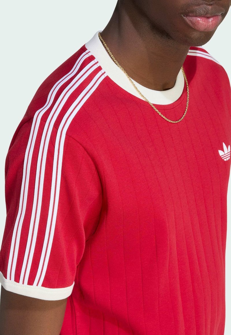 Homme portant un t-shirt rouge Adidas avec des bandes blanches sur les manches et le col, avec un petit logo sur la poitrine et une fine chaîne en or autour du cou.