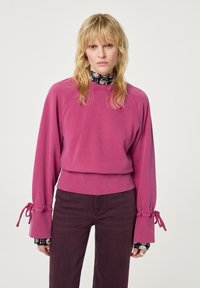 Fuchsia sweatshirt met lange, opbollende mouwen en verstelbare banden bij de polsen. Draagt over een zwart patroon top, gecombineerd met donkere broeken.