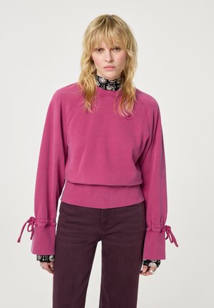 Fuchsia Sweatshirt mit langen, gepufften Ärmeln, die verstellbare Bindebänder an den Handgelenken haben. Trägt man über einem schwarzen gemusterten Oberteil, kombiniert mit dunklen Hosen.