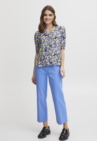 Blusa floreale con maniche a sbuffo corte in viola, giallo e blu, abbinata a pantaloni a gamba wide blu chiaro e scarpe nere.