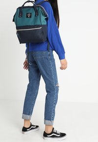 Personne portant un sweat-shirt bleu et un jean bleu déchiré avec les revers retroussés, des baskets noires et blanches, et un sac à dos bleu sarcelle et marine qui marche.