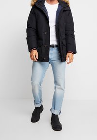 Sort isoleret jakke med pelsforet hætte, knapper foran, lommer, kombineret med lyseblå jeans og sorte ankelstøvler.