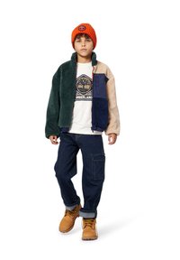 Flis jakna u zelenoj, bež i tamnoplavoj boji, nosi se preko bijelog majice s Timberland logom. Tamni traper cargo hlače i smeđe čizme upotpunjuju izgled.