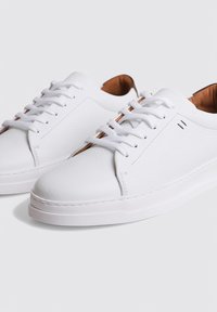 Baskets blanches en cuir avec une finition texturée, lacets plats blancs et doublure intérieure contrastante marron. Présentent des coutures minimales et une semelle en caoutchouc.