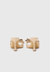 Nude høyhælte sandaler med et strømlinjeformet design, åpen bakside og fremtredende sløyfedetalj på overdelen. Tynn hæl med en svak gradient ved basen.