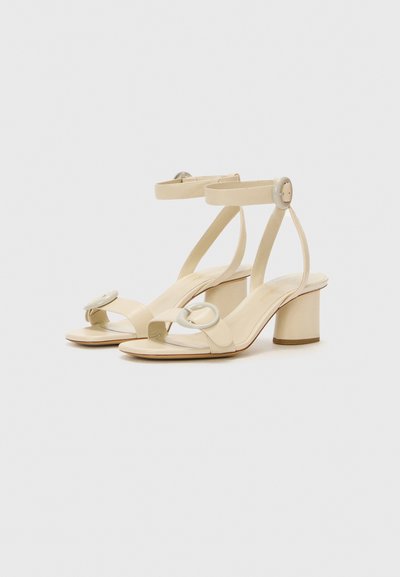 Beige højhælede sandaler med en glat finish, der har et dobbeltslyngedesign, cirkulære hardwaredetaljer og en kraftig hæl.