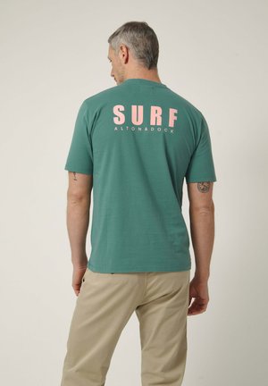 Hombre con cabello gris de espaldas, vistiendo camiseta verde con las palabras "SURF ALTONADOCK" en letras rosas y pantalones beige sobre fondo liso.